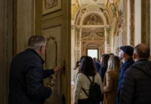Le Vie dei Tesori compie vent’anni: i luoghi dell’edizione 2026 Una guida apre una porta affrescata a un gruppo di visitatori dentro un palazzo barocco.